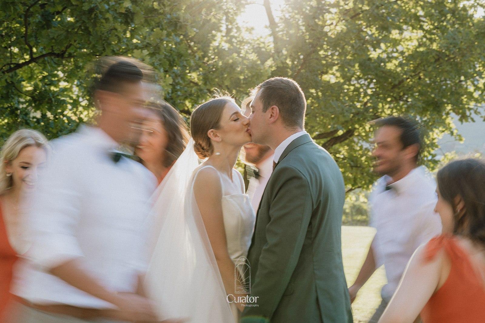 Wedding couple kiss with a motion blur of their wedding party running around them