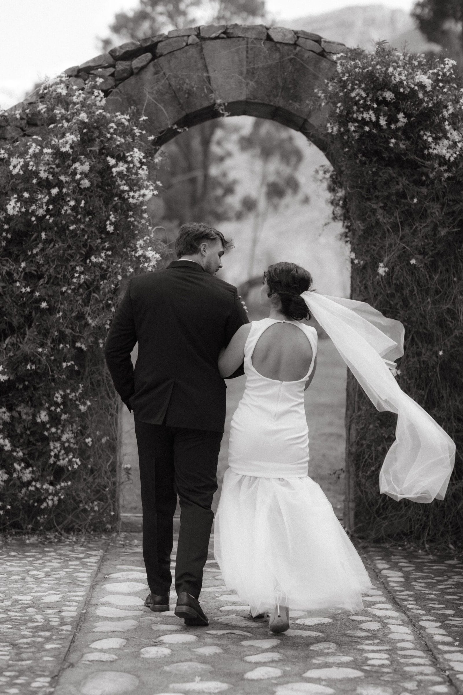 Black and white of a wedding couple walking towards a stone arch at La Roche Estate